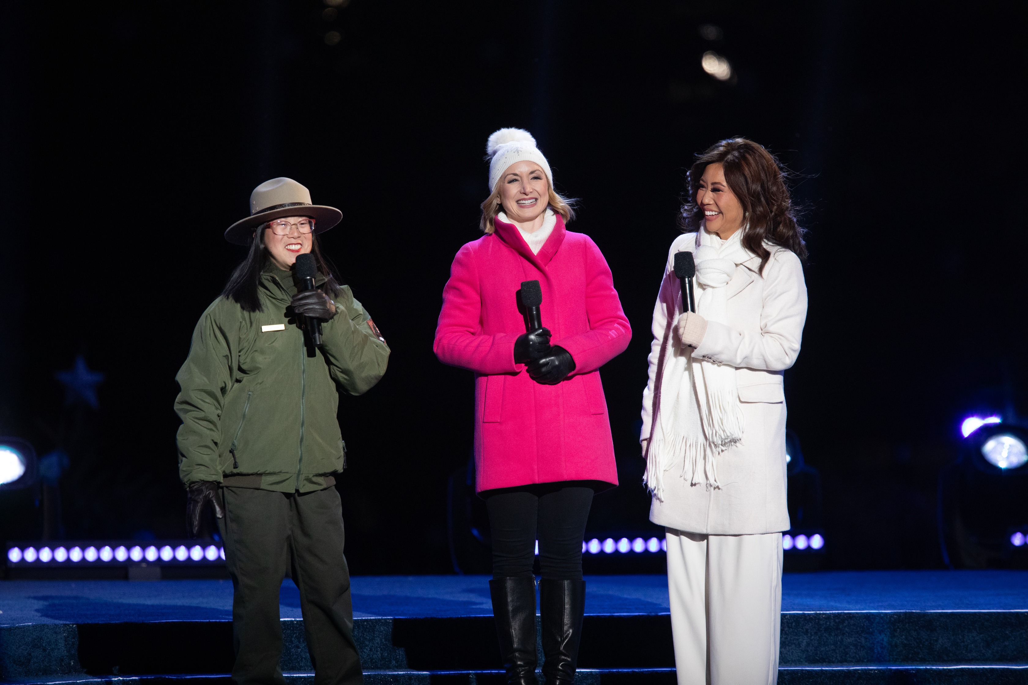 A park ranger and two women smile on a stage while they hold their microphones. 