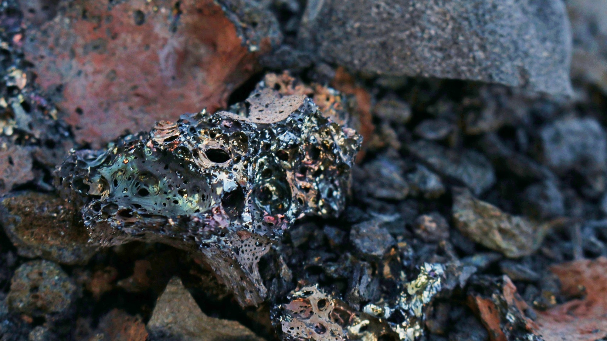 closeup of several small black and reddish stones with many air bubbles, three of the rocks have a shiny multicolored glaze on them