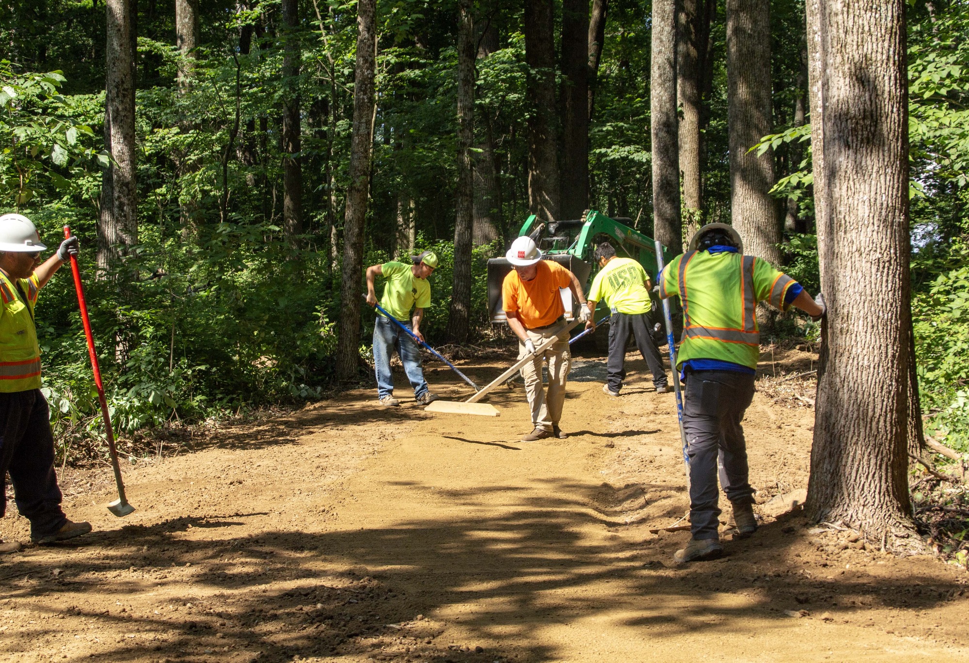 Five construction workers use rakes to smooth out the brown stabilized aggregate 	material with trees and a small green construction vehicle in the background.