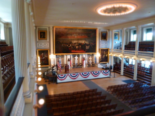 Interior view of Faneuil Hall, an open two-story meeting space with rows of wooden chairs in front of a stage. On the stage are chairs, a row of flags and busts. The space is flanked by two galleries with stacked rows of wooden chairs. Portraits of politicians and leaders are displayed on the walls.