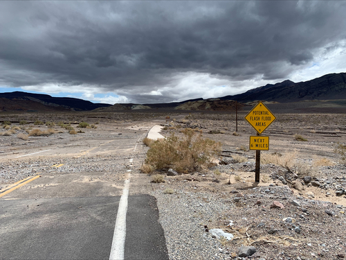Gravel and mud mostly cover a paved two-lane road in a desert landscape near a road sign which reads "Potential Flash Flood Area Next 6 Miles," with dark clouds overhead.