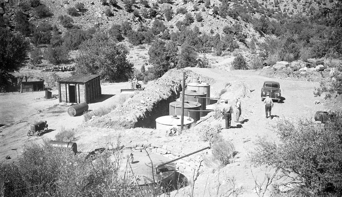 Men standing next to open trench with four underground storage tanks set in place in the Oak Creek Maintenance Area. Tanks were obtained from war surplus, used for storage of road oil. Installation is faulty in that the tanks were set too low for loading. Tanks will have to be raised about three feet to facilitate loading out. Machinery and structures in background.