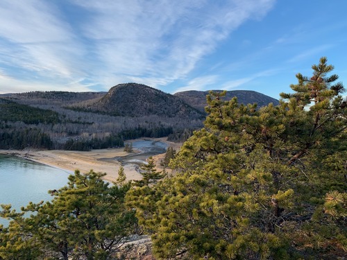 Green trees in foreground, sand beach below and mountains in back. Ocean is on the left side of the frame and blue sky above. 