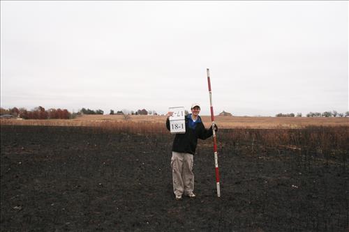 A person standing in the prairie with a stadia rod and a sign.