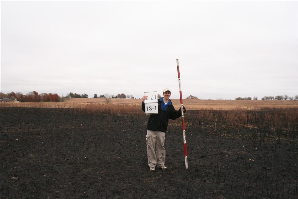 A person standing in the prairie with a stadia rod and a sign.