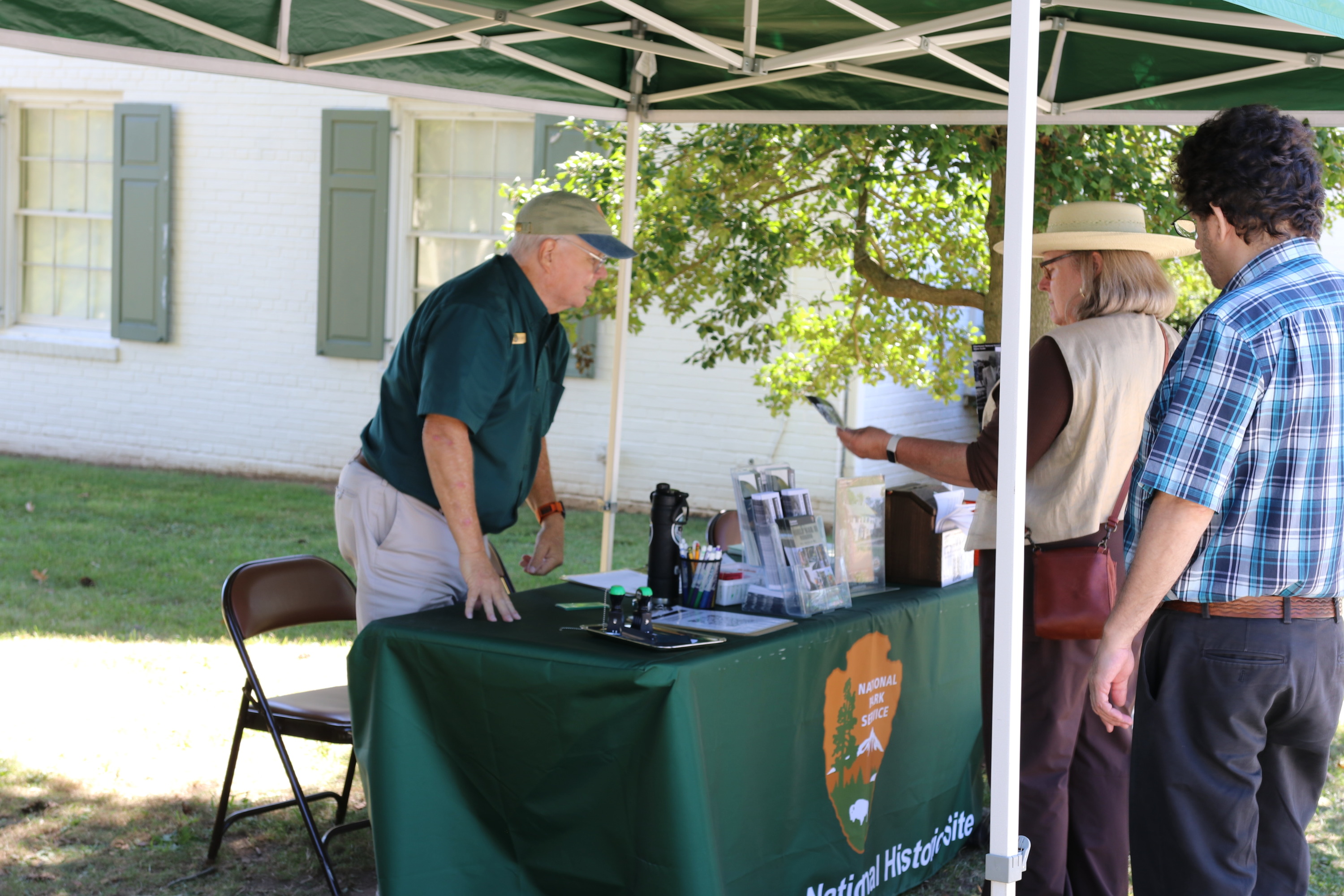 A man in a green shirt speaks with visitors under a green tent. 