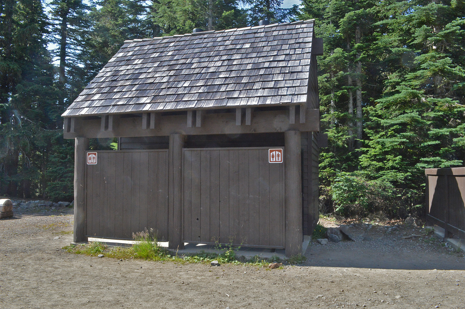 A small brown restroom building with steep, shingled roof is surrounded by hard packed gravel. A forest fills the background.