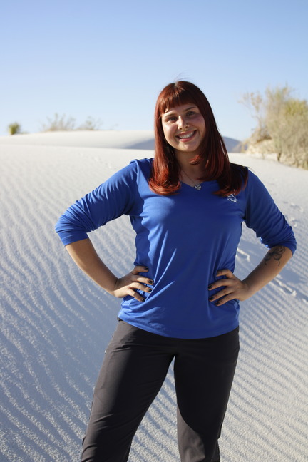 A young woman stands for a photo on rippling white sand dunes. Behind her desert shrubbery grows from the sand and the dunes are met by a clear blue sky. 