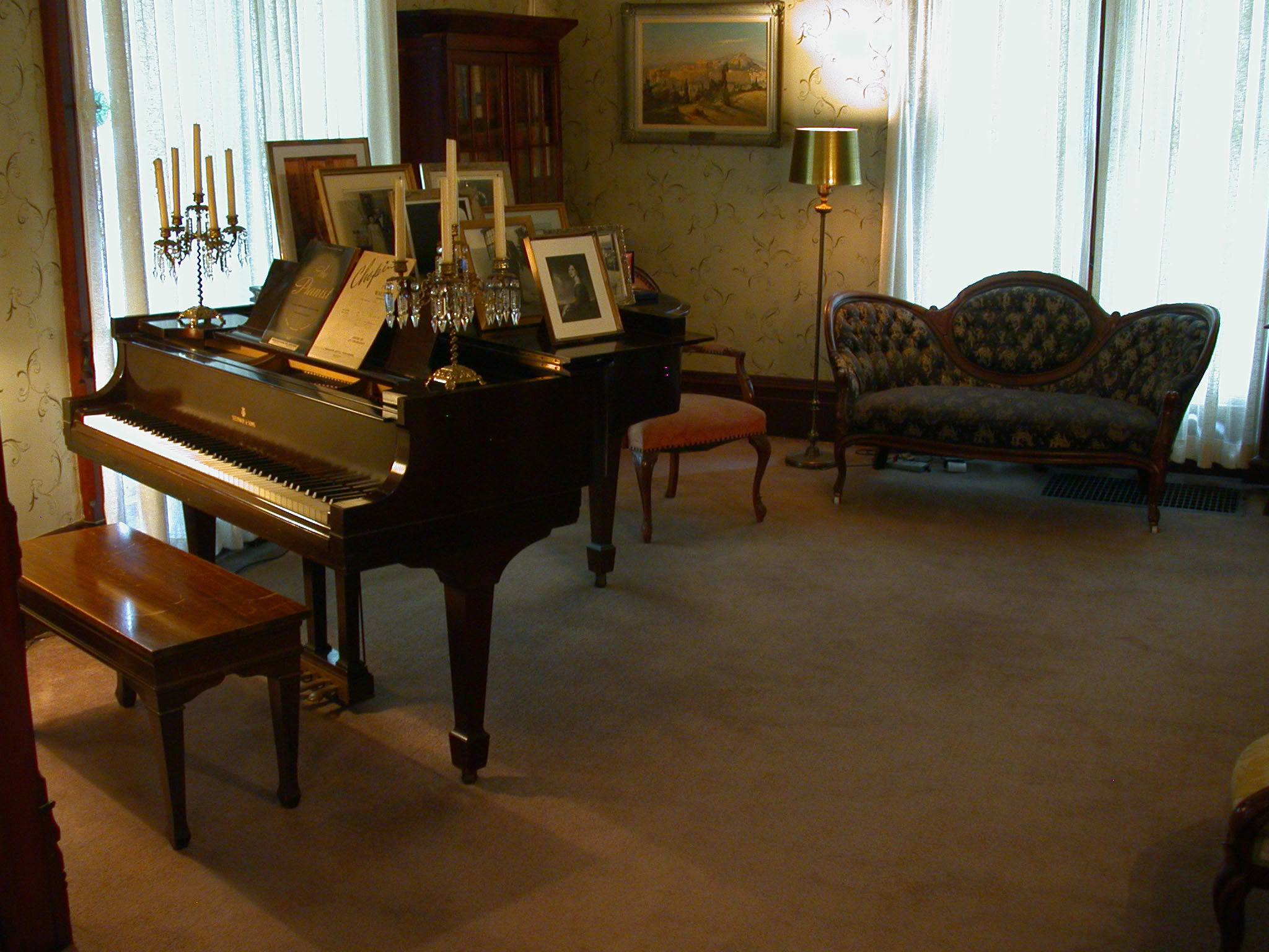A color photograph of the music room/ parlor. A baby grand piano is adorned with candelabras and photographs. An antique sofa and chair are prominent. 