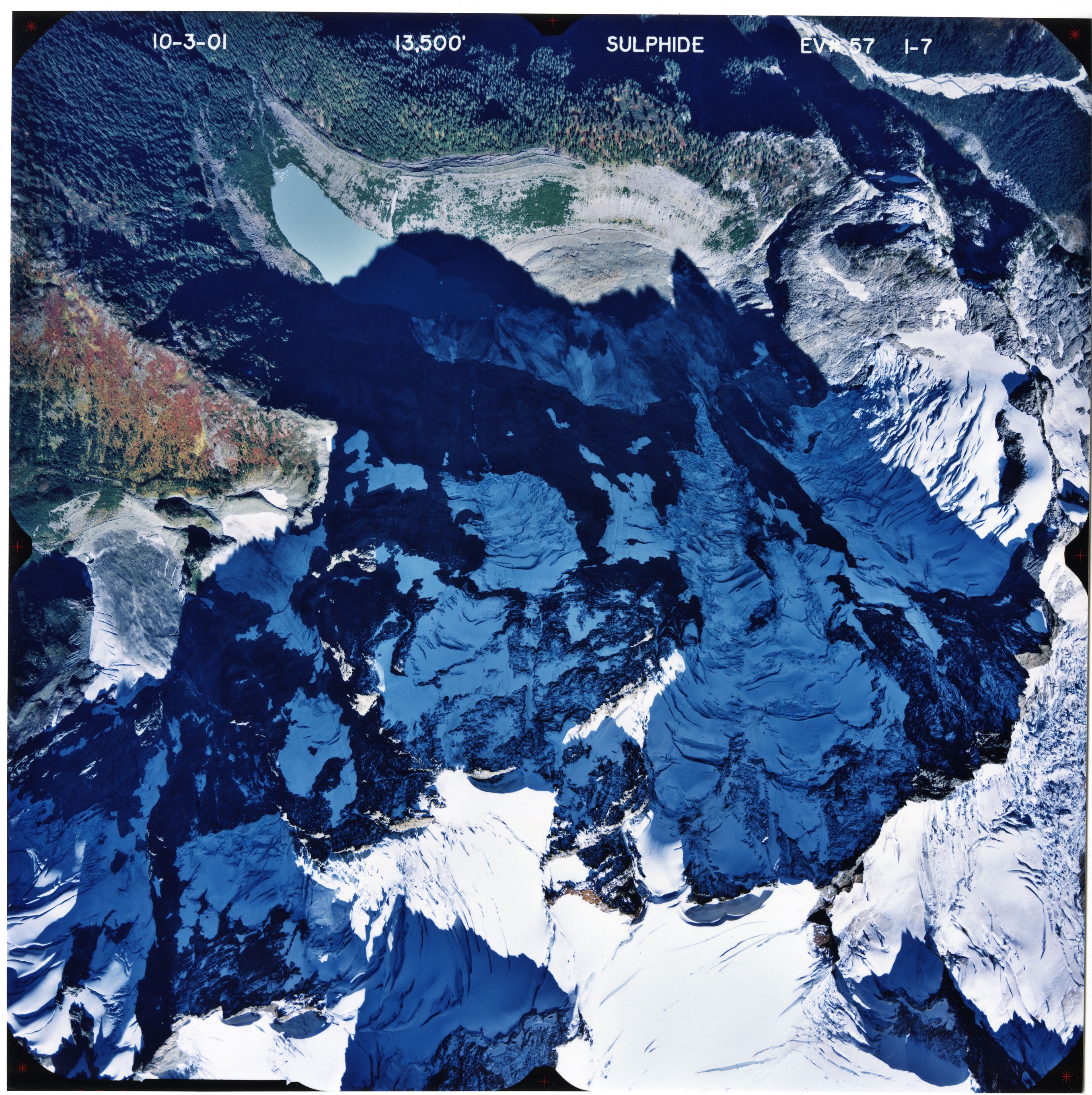 Color aerial photograph of glacier on mountain peak with forested valleys below.