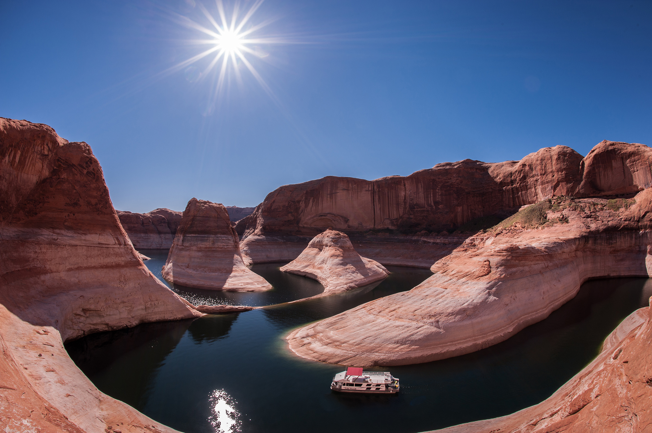  Photograph of Reflection Canyon in Lake Powell with a houseboat navigating the curves of entrenched meanders among steep cliffs. 
