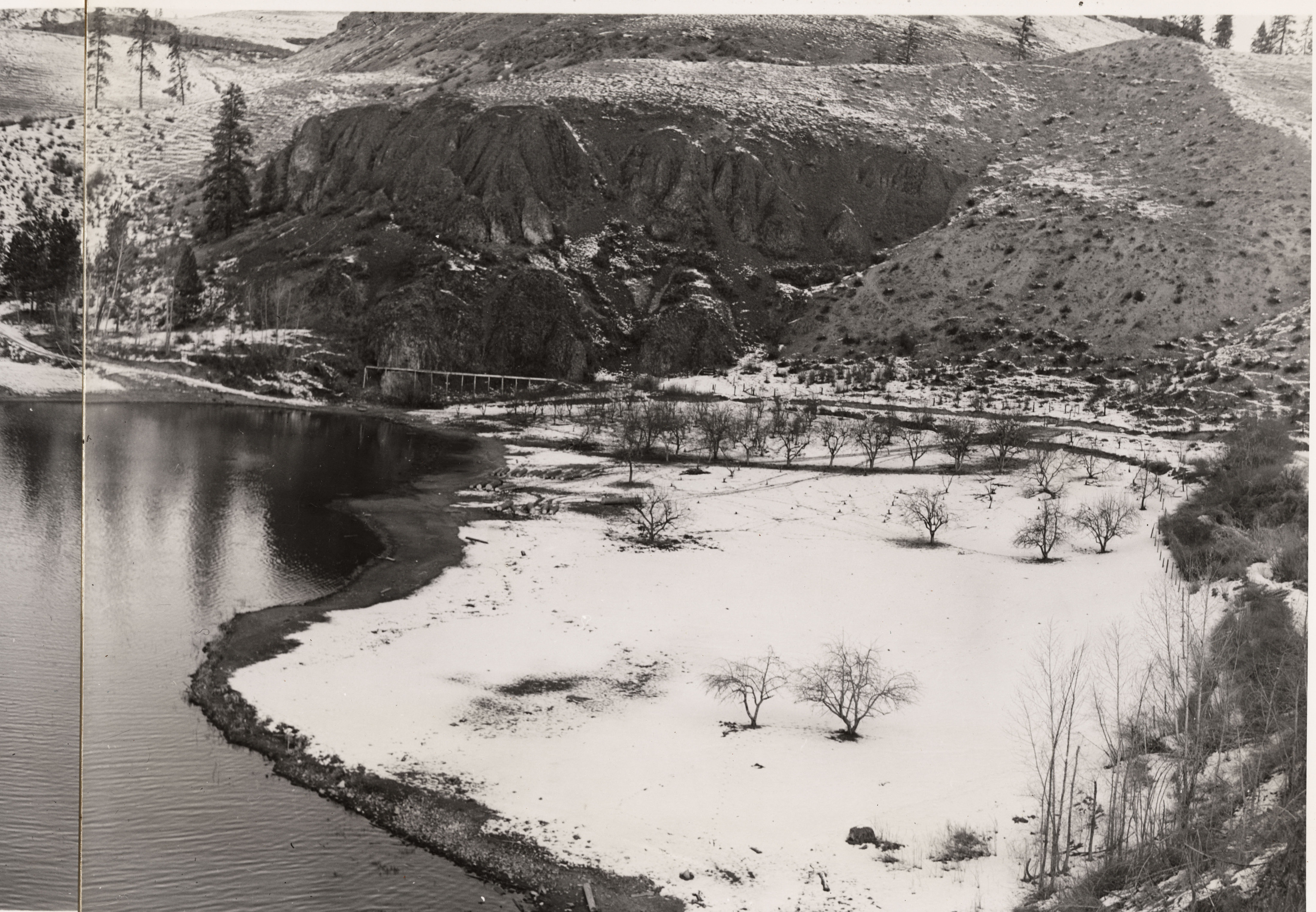Black and white photograph of the snowy shoreline of a body of water with steep, dark cliffs in the background