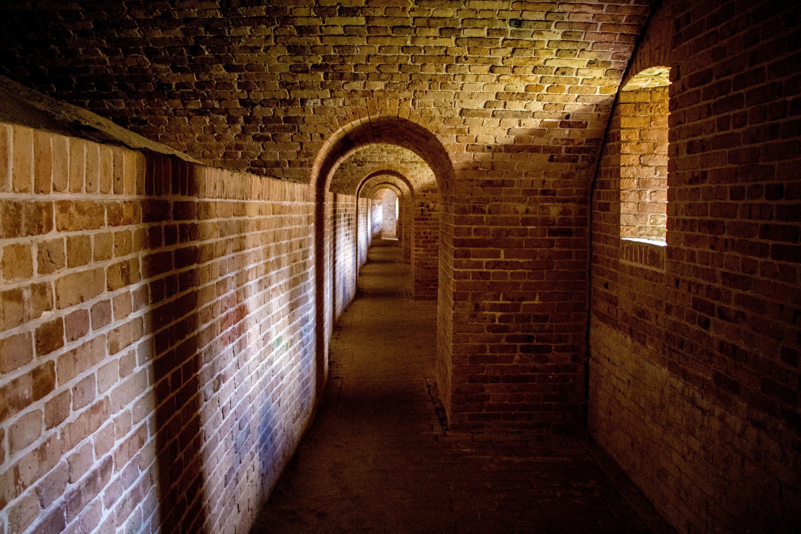 The sun shines through small loopholes (windows) into a brick arched gallery.