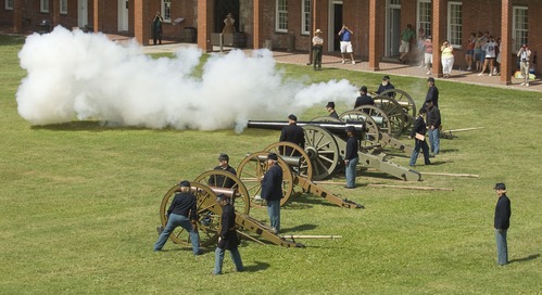 Four cannons lined up in a row with people in Civil War Period dress around. The furthest cannon has smoke coming from it. 