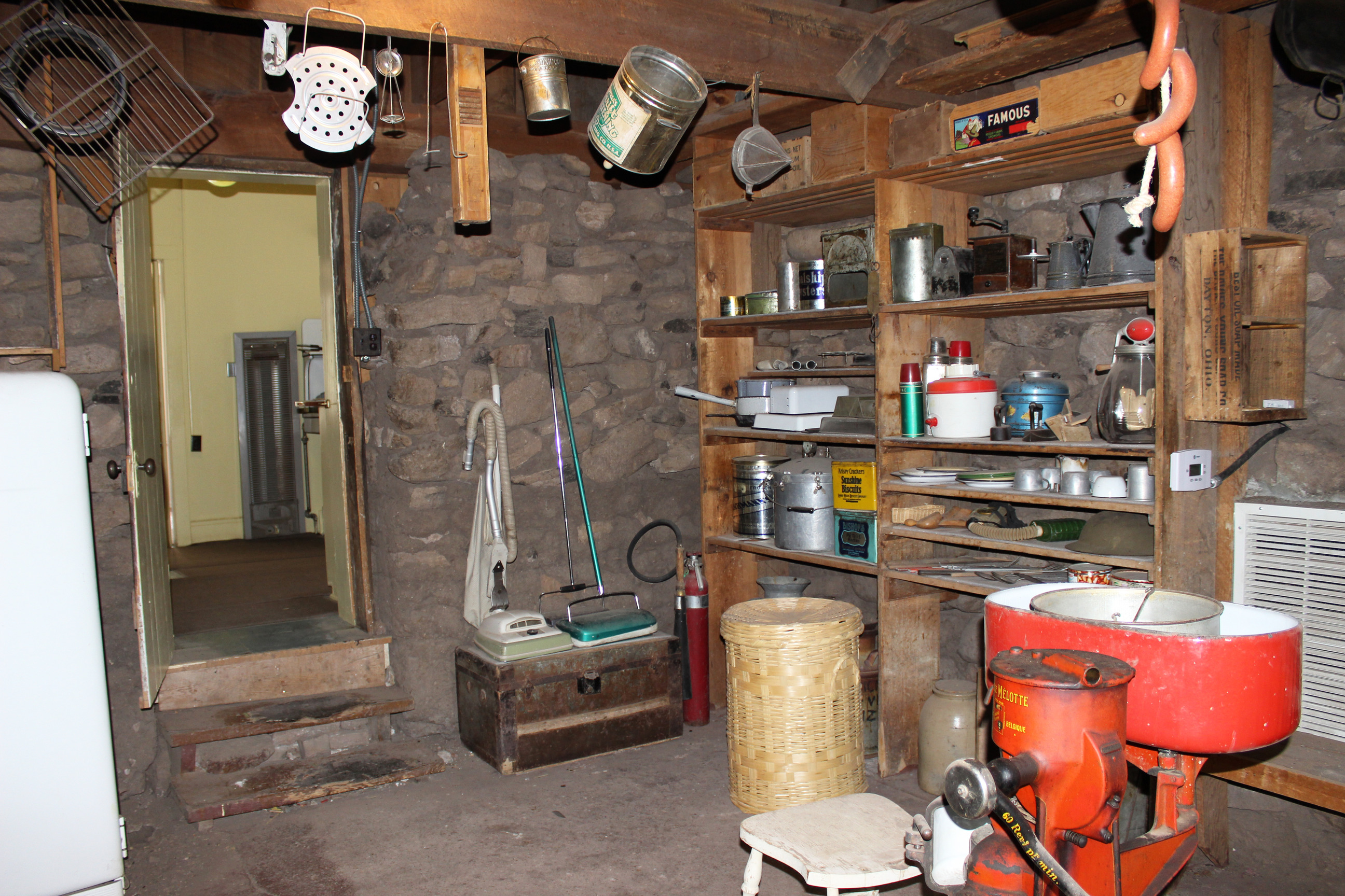 Cellar with rough stone walls, wooden shelves, miscellaneous household and kitchen items, including a red milk and cream separator. 