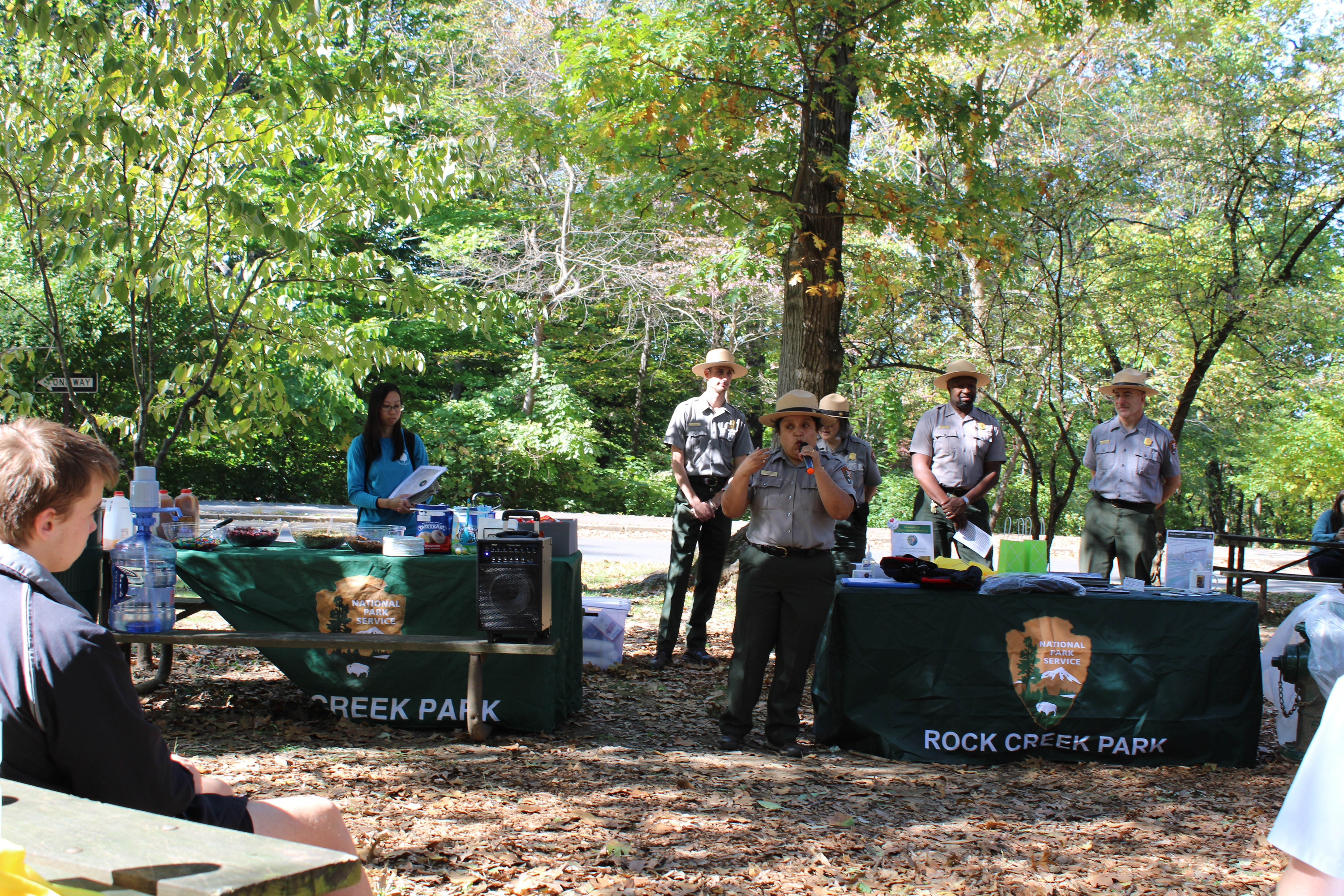 Standing in front of two National Park Service branded tables, a NPS uniformed woman speaks into a microphone. A crowd of people sitting at picnic tables watch and listen.