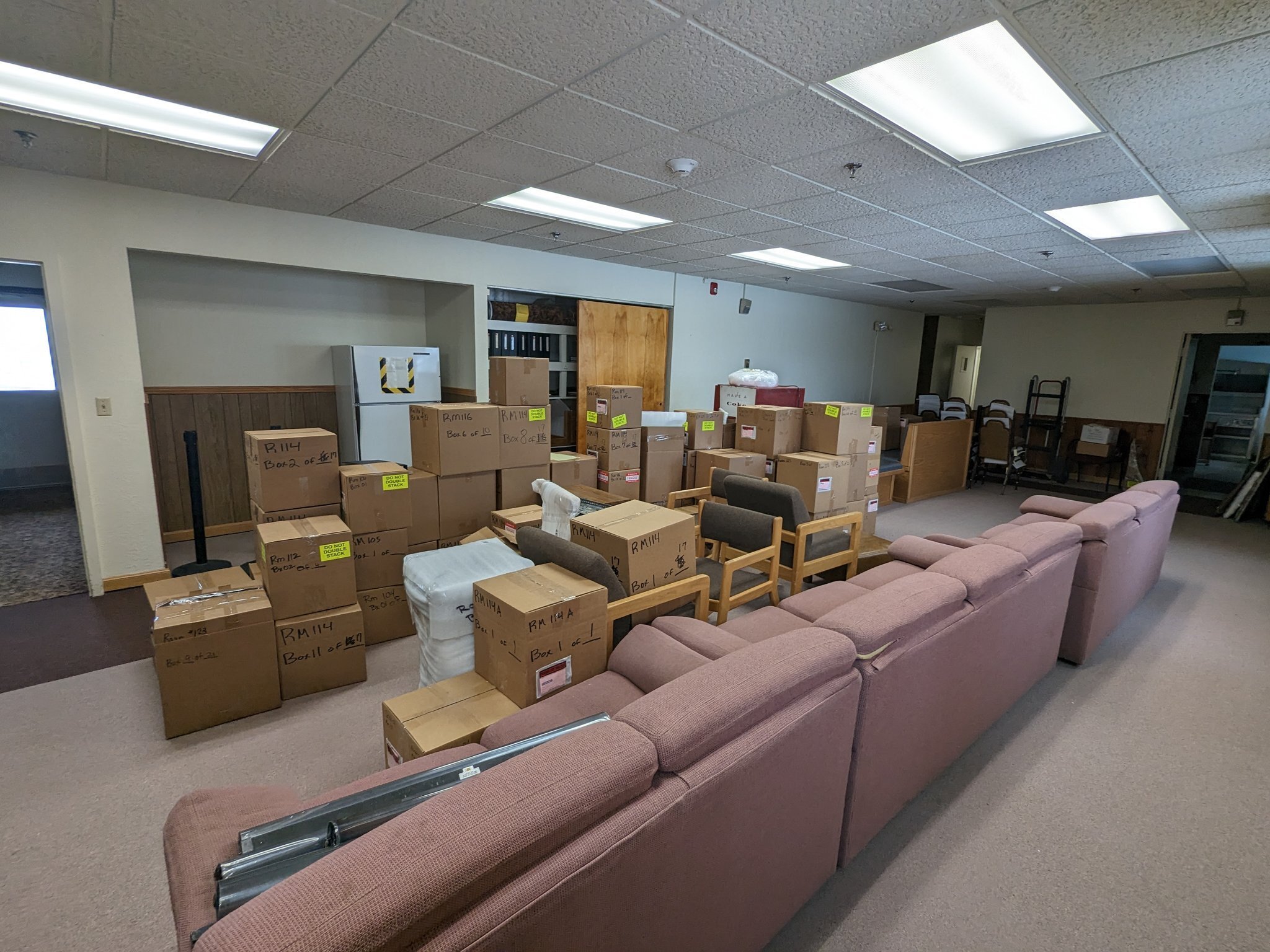 A long room with white walls and wood paneling. Stacks of boxes are all over the floor, some fabric couches, some chairs, a refrigerator, and an old coke machine.