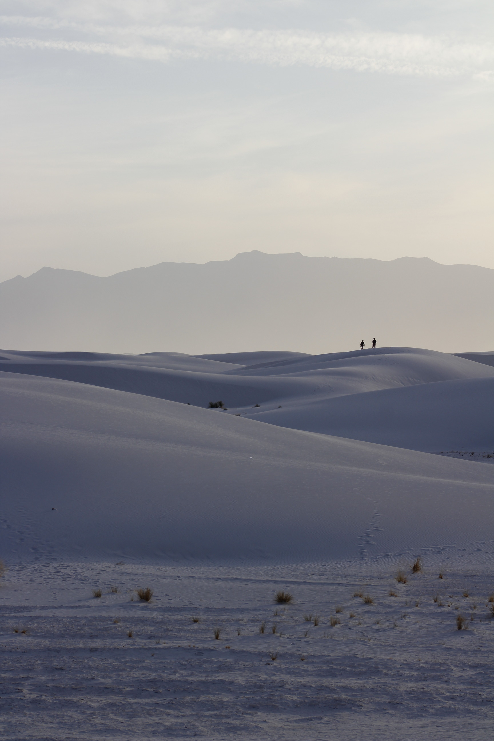 Two people stand on a sand dune, in the distance rolling white dunes meet mountains rising on the horizon. 