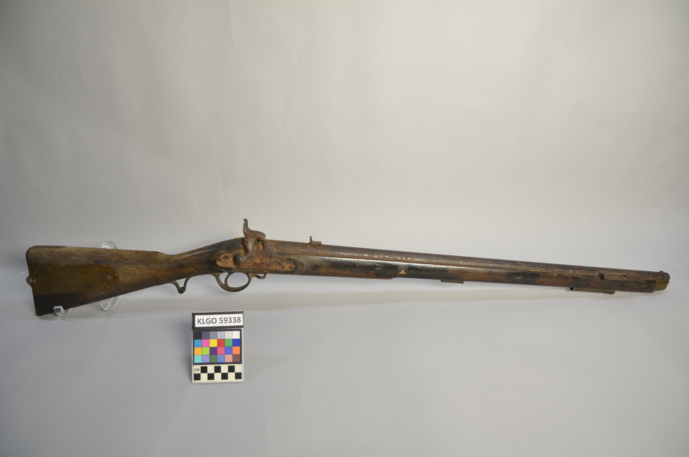 a color photo of a rifle with rusted barrel and light brown stock and a hinged brass plate on butt and right side