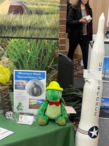 Table display in a hotel lobby. Table has a green cloth, a plush turtle with yellow helmet, and a large white model of a missile. 