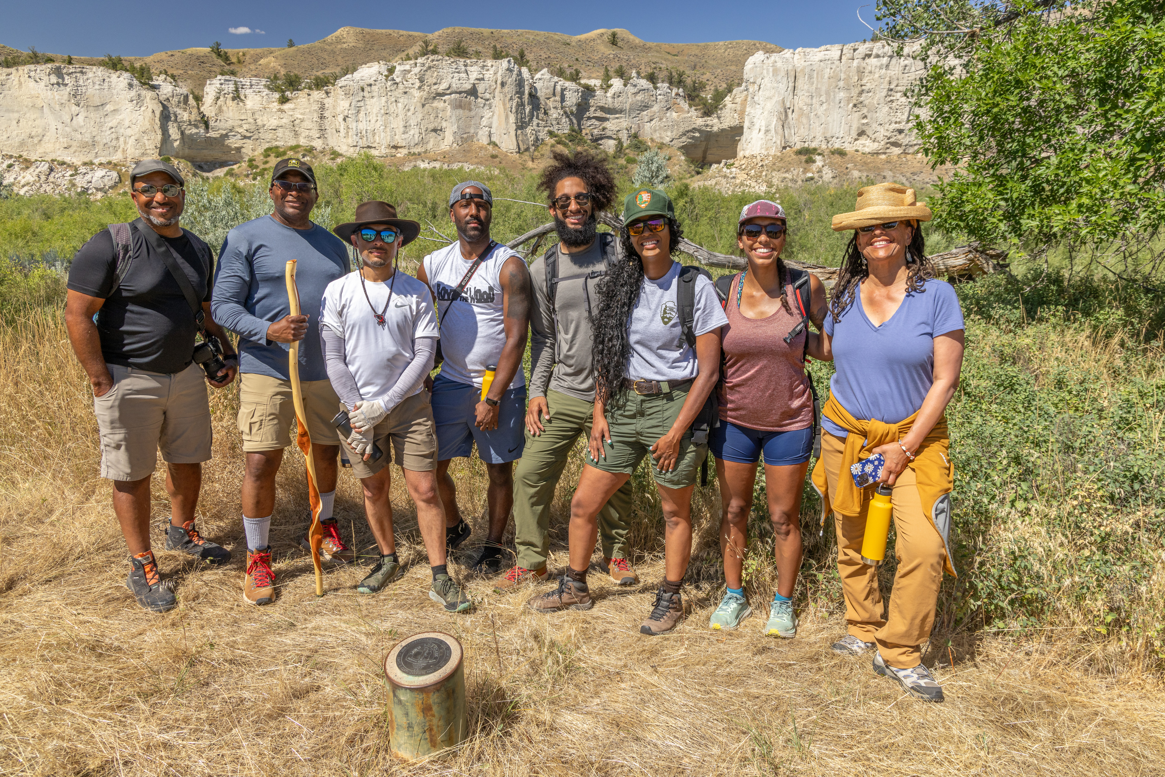 Eight people posing in front of a small, cylindrical Lewis and Clark campsite marker with rugged scenery in background 