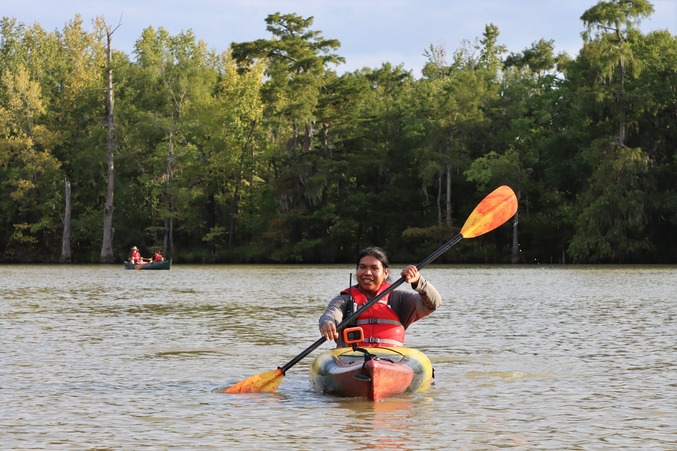 a man paddling a red-yellow-black kayak with black and orange paddles on a wide waterway lined with tall trees.