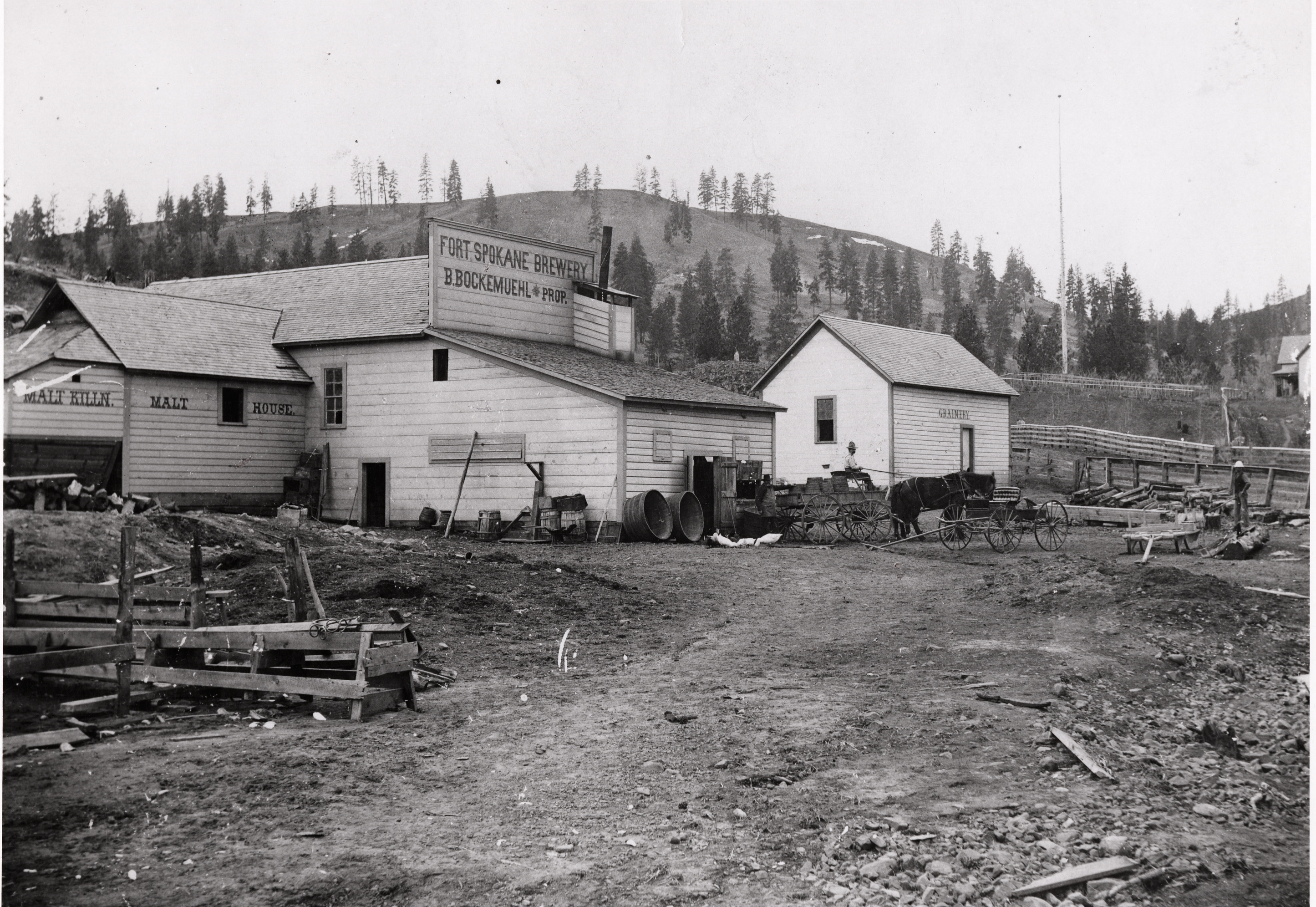 Black and white photograph of plain wooden building with a small building next to it and horses and wagons in front