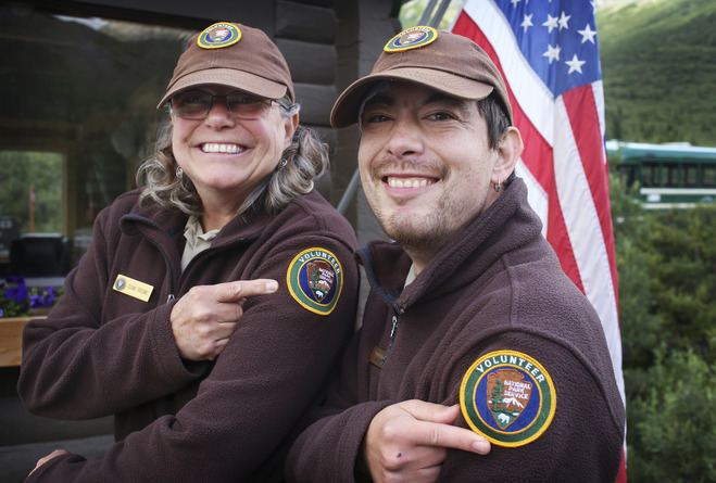 a man and woman in national park service volunteer uniforms smile and point to volunteer patches on their shoulders
