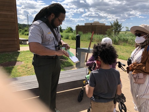 On the left side of the picture, a park ranger holding a clipboard can be seen explaining the prompts in an activity sheet to a young boy, and two older women. All three are standing on tan pavement. A gray wooden bench can be seen behind the group. A small building, tall grass, and large trees can be seen in the background.