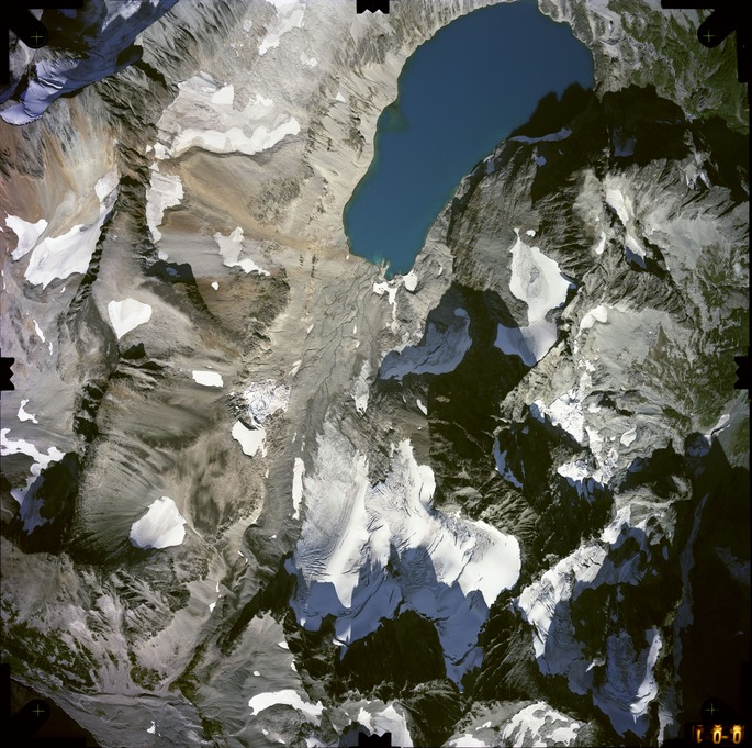 Color aerial photograph of glacier, snowfields, and alpine lake on mountain peak with forested valleys below.