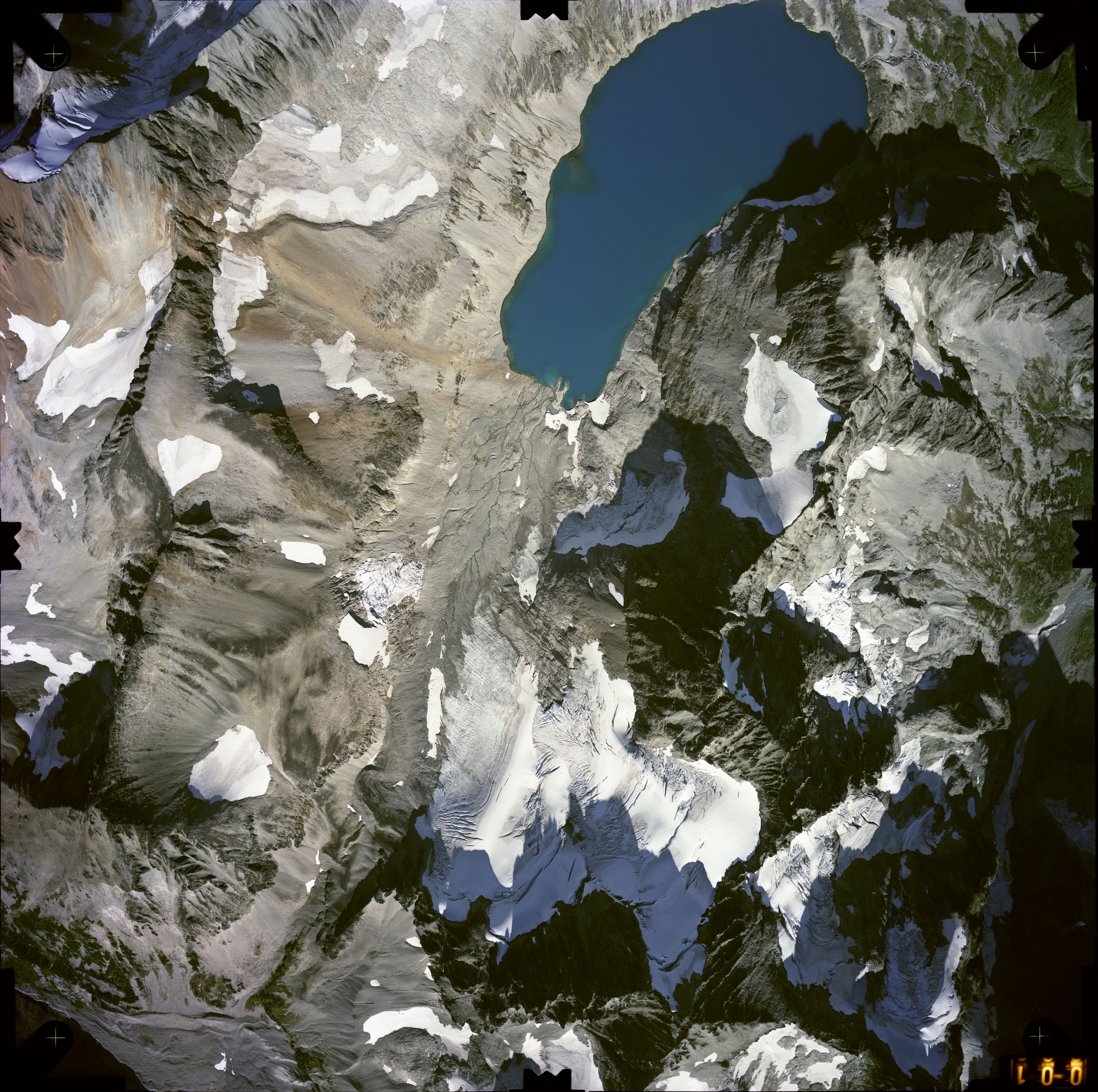 Color aerial photograph of glacier, snowfields, and alpine lake on mountain peak with forested valleys below.