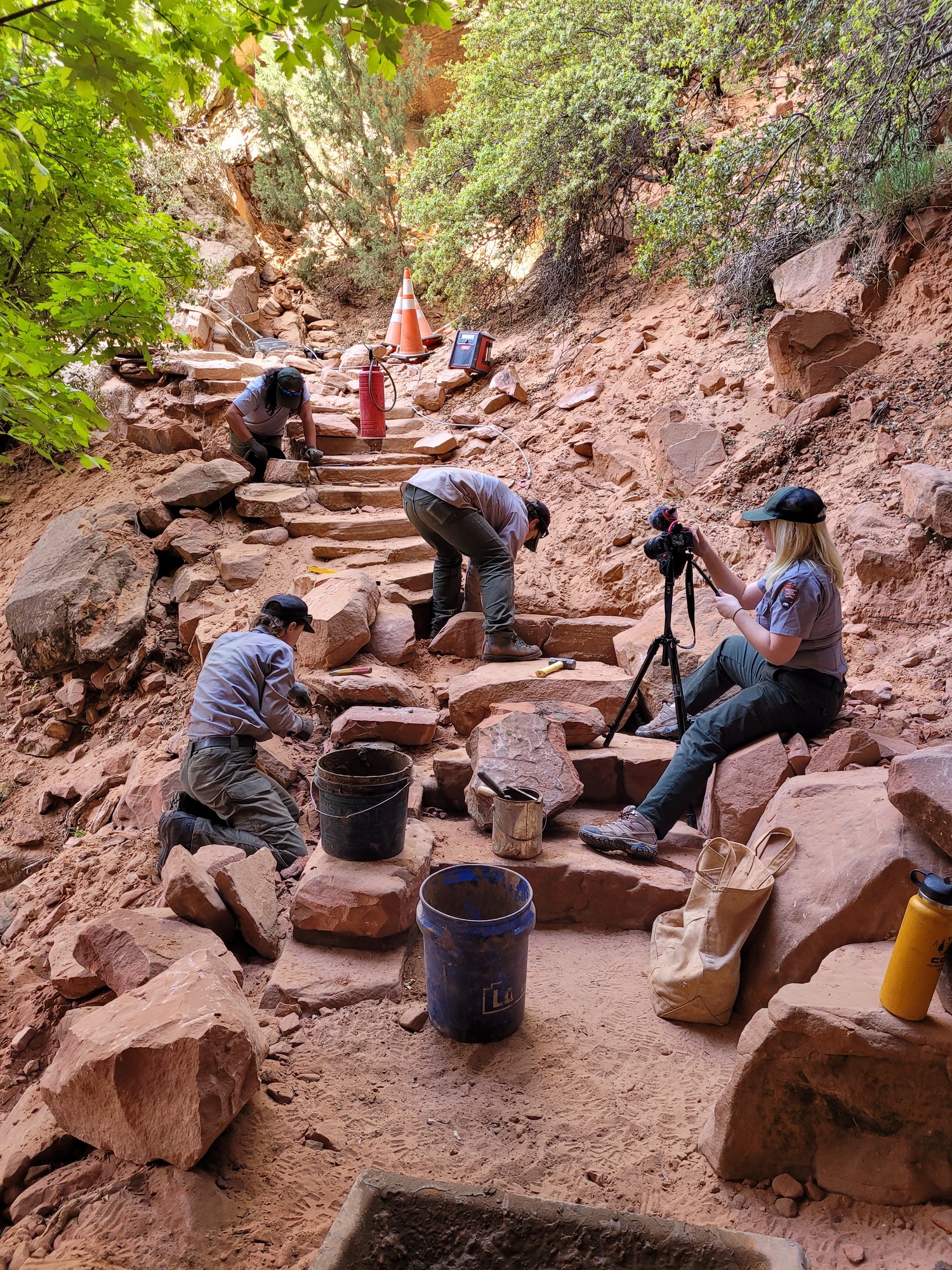 Four rangers sit on stone stairs. Three of the rangers are repairing the stairs, and the fourth is capturing video of the work. 