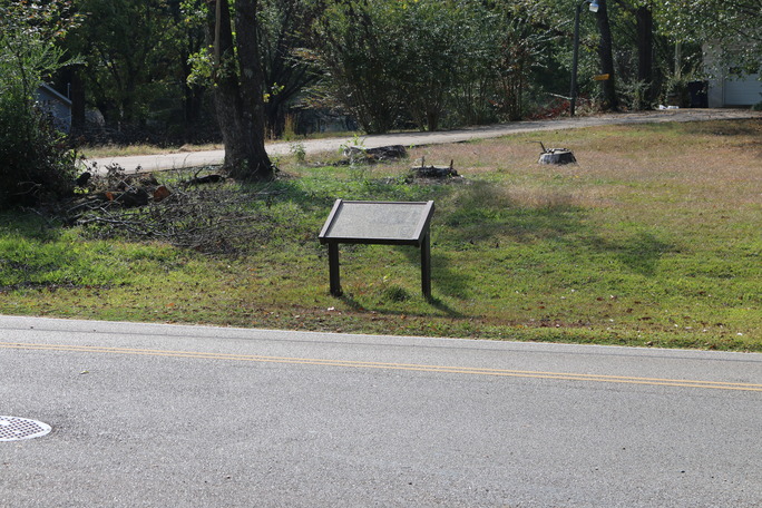 A battered wayside stands in a front yard alongside a road. Trees are in the background.