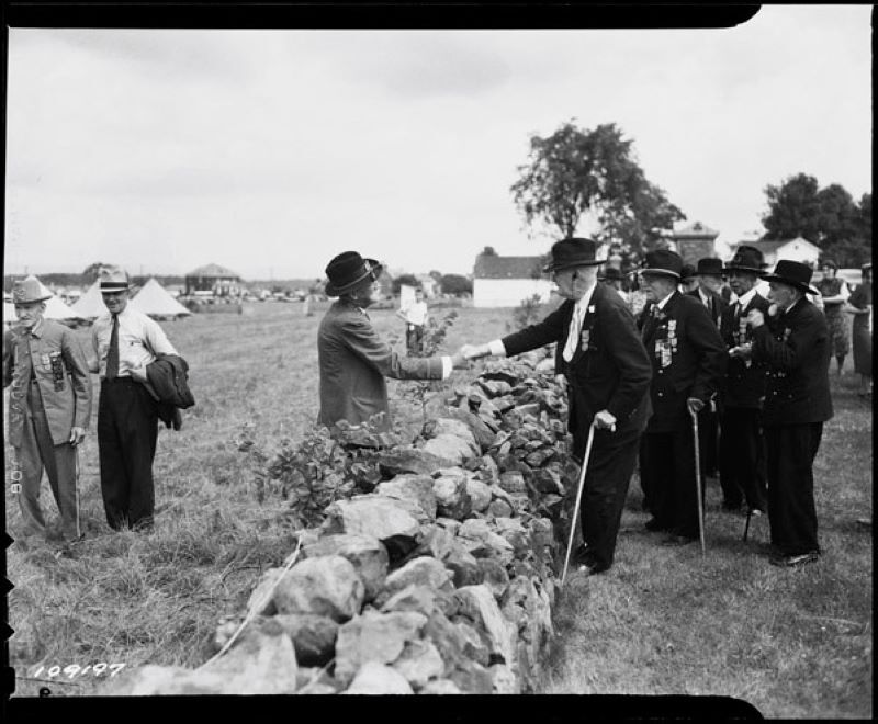 Photograph of Union and Confederate veterans shaking hands across a stone wall at the 1938 "Blue and Gray Reunion" at Gettysburg. Three men are on the left of the wall. Six men are to the right of the wall. Two men shake hands.