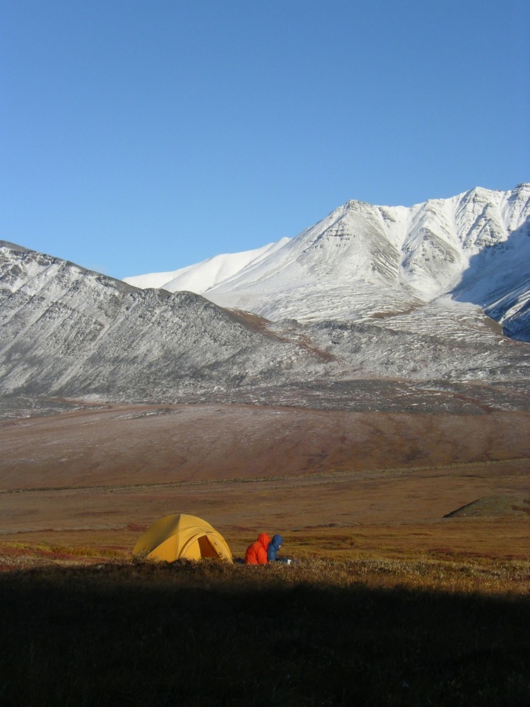Two wilderness travelers sit on the tundra outside their tent enjoying the midnight sun.