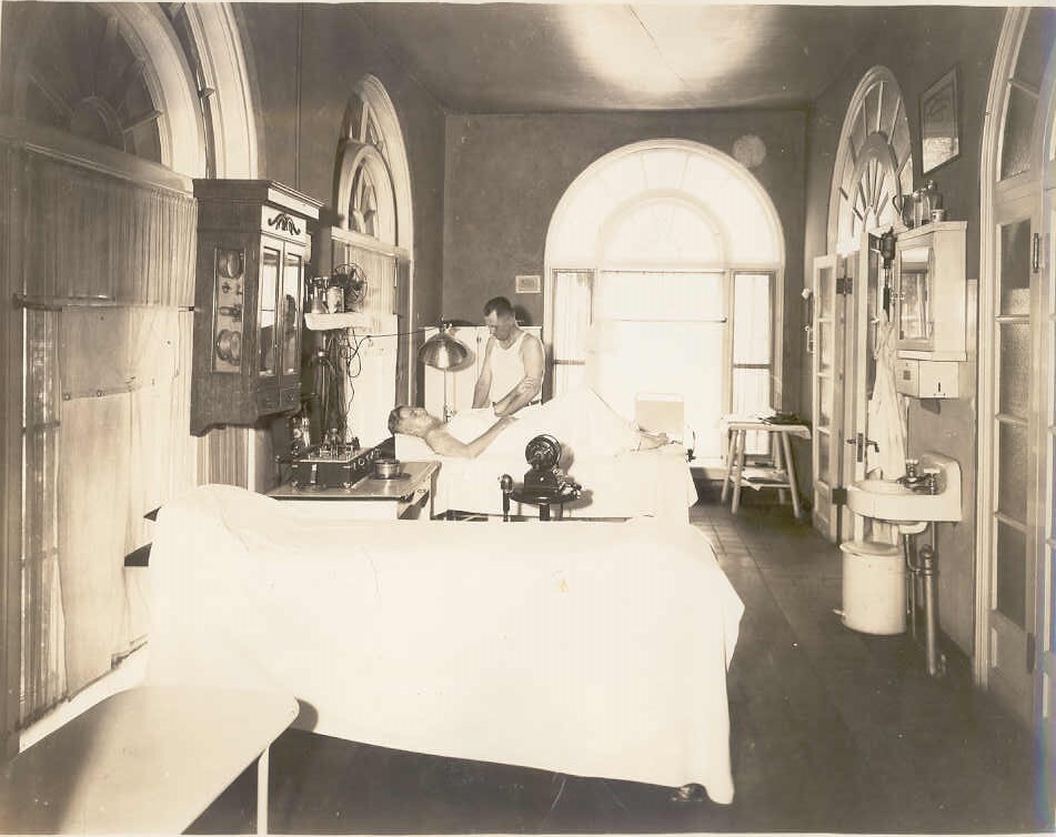 A masseur treating a male patron to a massage. Massage tables and other equipment are visible around the room. 