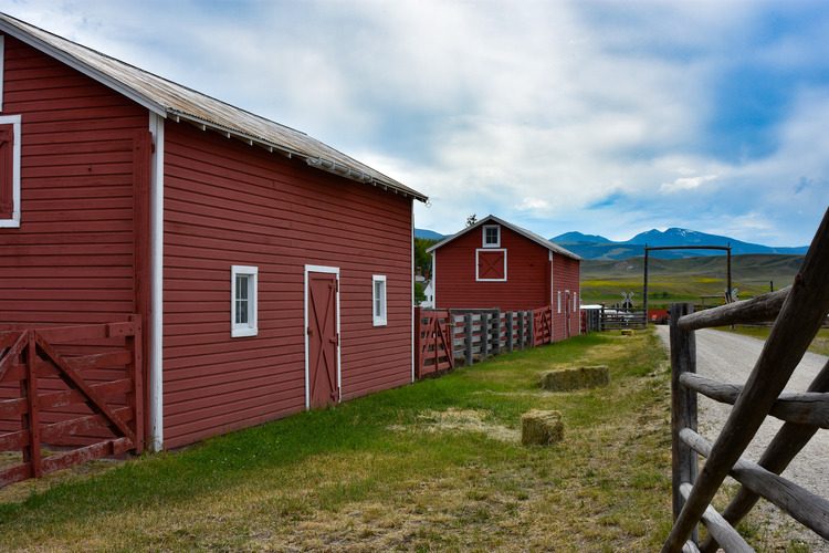 Two rectangular structures with siding and gable roofs, in line with a five-rail wooden fence. A gravel drive is parallel to the fence, leading through a gate to a view of open ranch land and mountains beyond. 