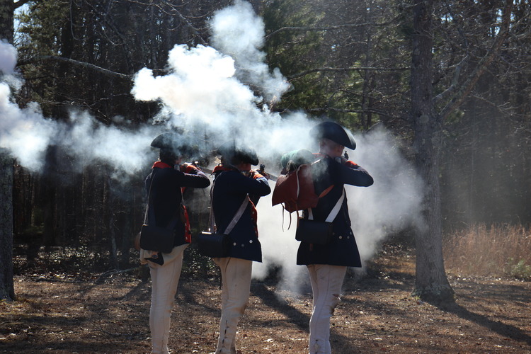 Three people dressed as Continental soldiers fire muskets in a cloud of smoke towards the woods. 