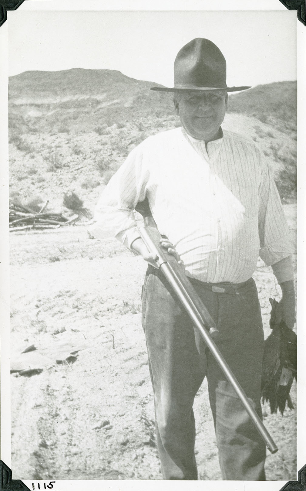 This is an historic black and white photograph from the Scotty's Castle Historic Photograph Collection, Death Valley National Park of heavy set, middle-aged man holding shot gun under right arm and dead ducks by neck in left hand. Wearing tall, peaked cowboy hat, white stripped shirt, and jeans. Standing in a desert environment with colored butte in background. Number in black ink in lower left corner.