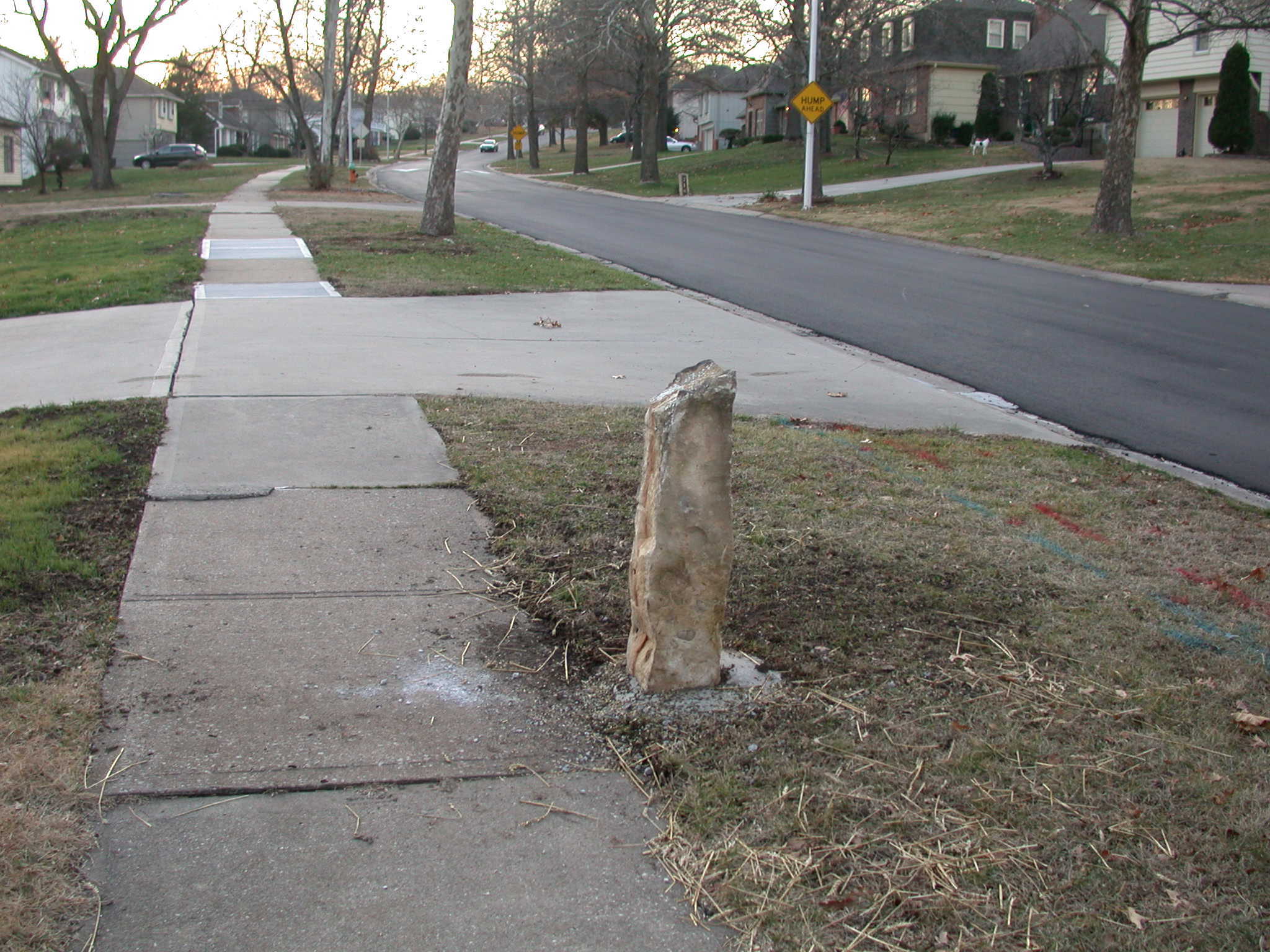 A weathered concrete post beside a sidewalk with a road and houses in the distance.