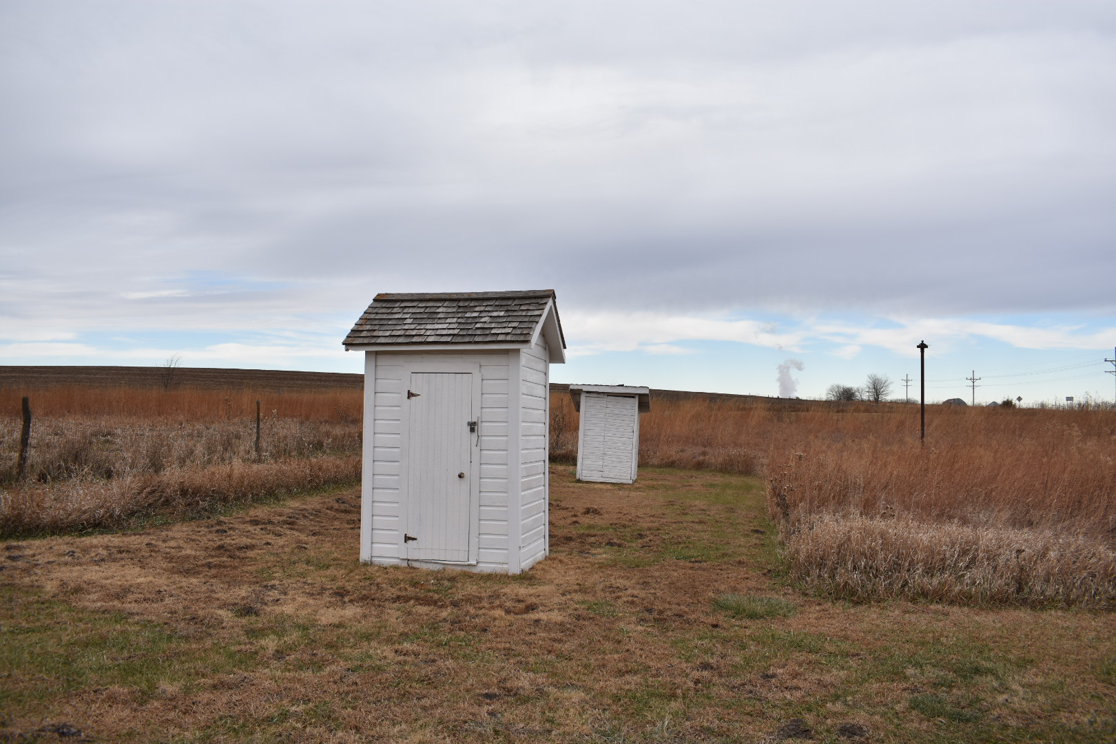 Two privies with white siding stand near each other in an area of mowed grass, with tallgrass prairie beyond.