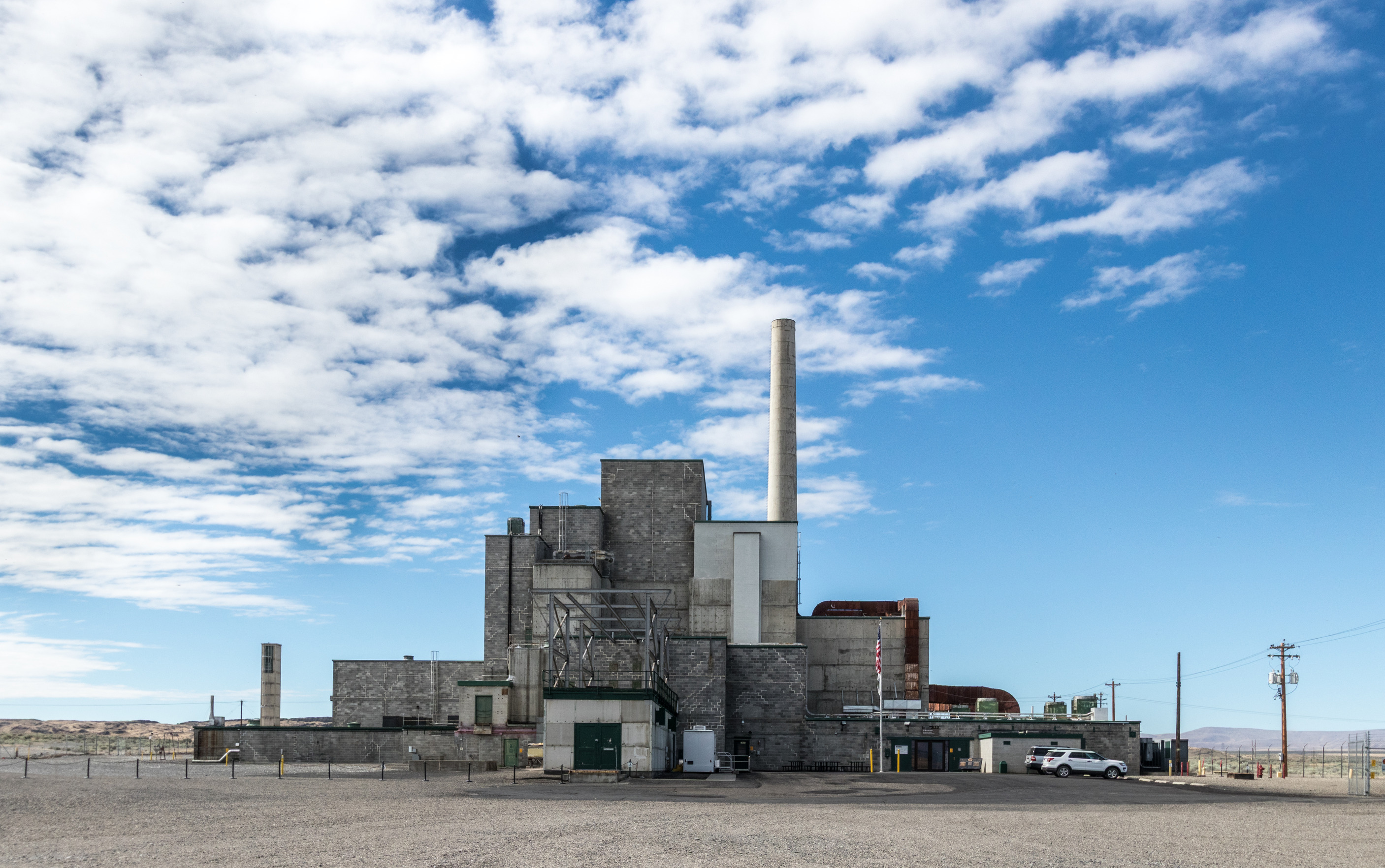 The B Reactor: A large building and gravel lot constructed in a stacked pyramid shape with a single ventilation stack standing taller than the building. Blue sky with white clouds above.