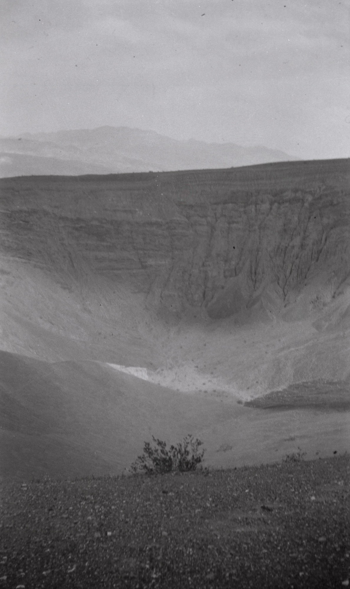This is an historic black and white photograph from the Scotty's Castle Historic Photograph Collection, Death Valley National Park of a large, volcanic crater. Gravel foreground with short desert plant. Far wall showing sedimentary layers and vertical erosion. Higher elevation mountain shows faintly in background.
