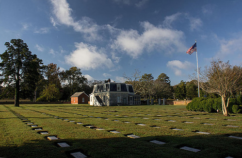 Rows of grave markers are flush with short turf in a cemetery. In the rear center, a lodge and smaller brick shed stand near a brick perimeter wall. A flag and several trees and shrubs stand in the cemetery. 