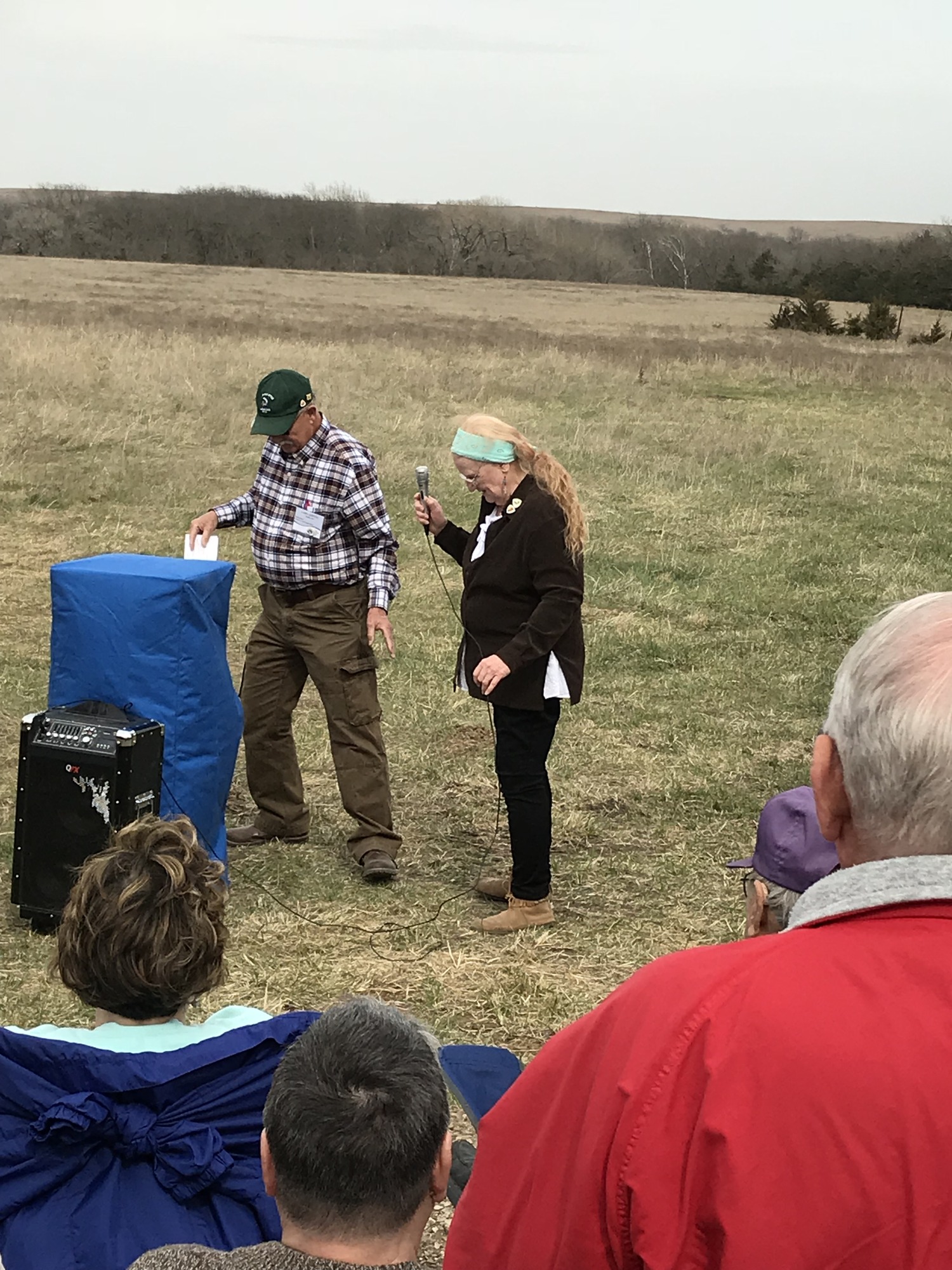 Two people holding microphones are presenting in front of a small outdoor audience with grassy fields in the background.