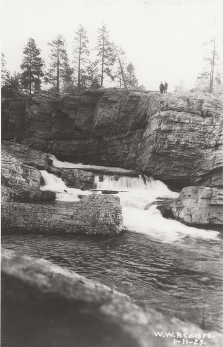 Black and white photograph of two men standing on a rock cliff above a waterfall