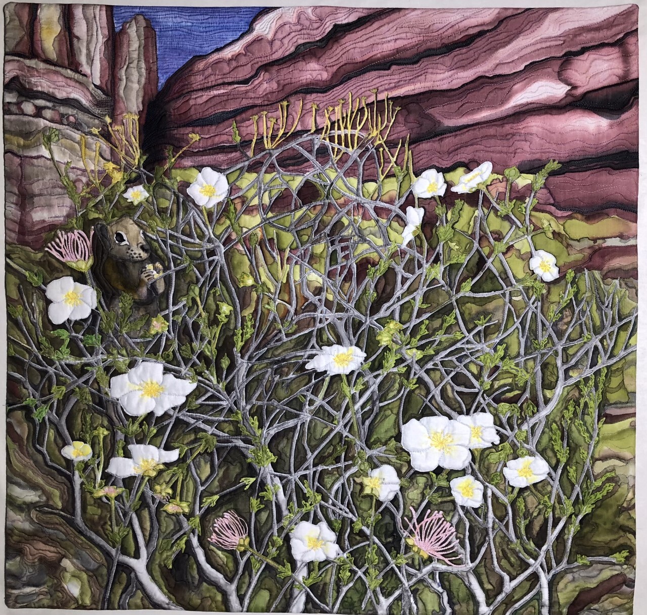 Quilted and painted fabric creates a scene. A woody shrub with white flowers grows among tall purple cliffs.
