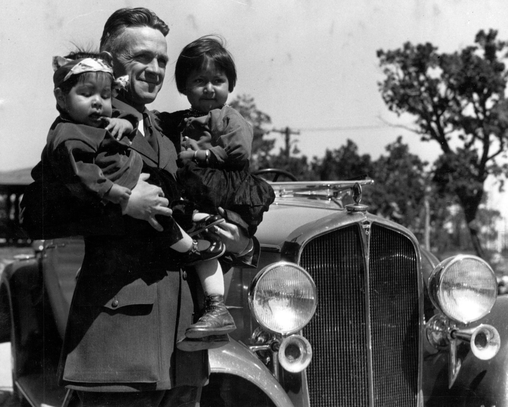Black and white image of Superintendent Tillotson holding Porter Timeche's young children while standing next to an old vehicle.