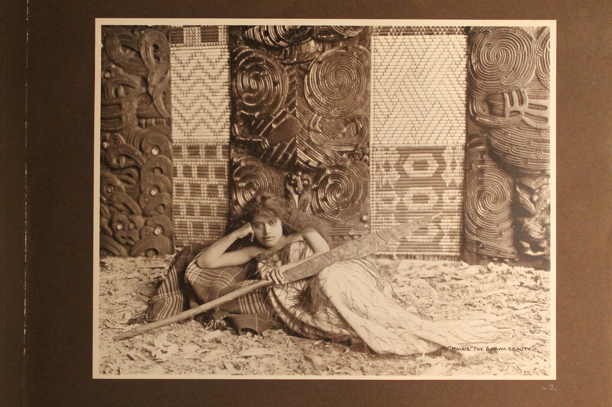 A Maori girl reclines on a plaid blanket within an elaborately carved interior.