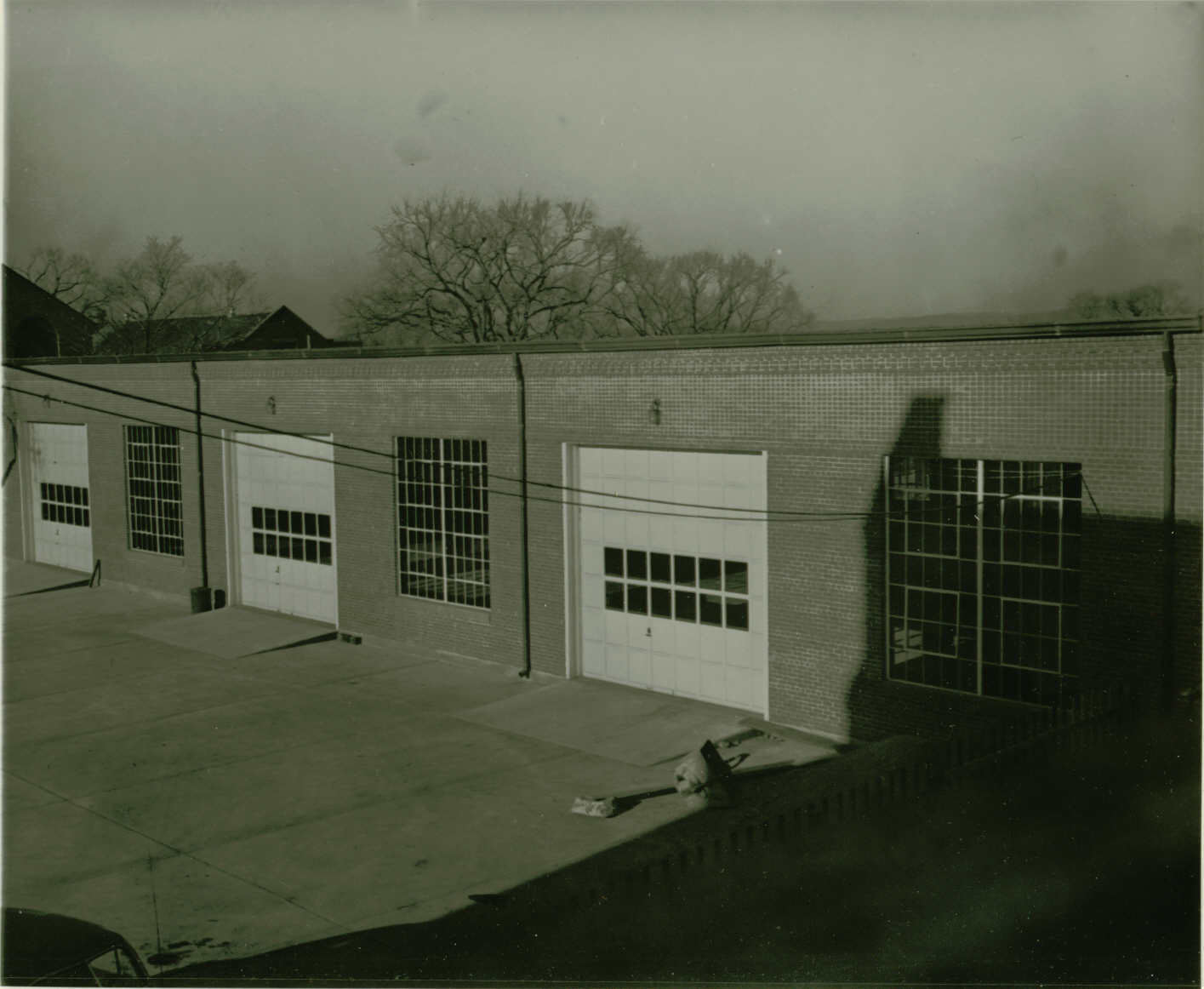 Black and white photo taken from above of a brick building with three closed roller doors. Concrete ramps lead up to those doors. Large square windows sit between the doors.